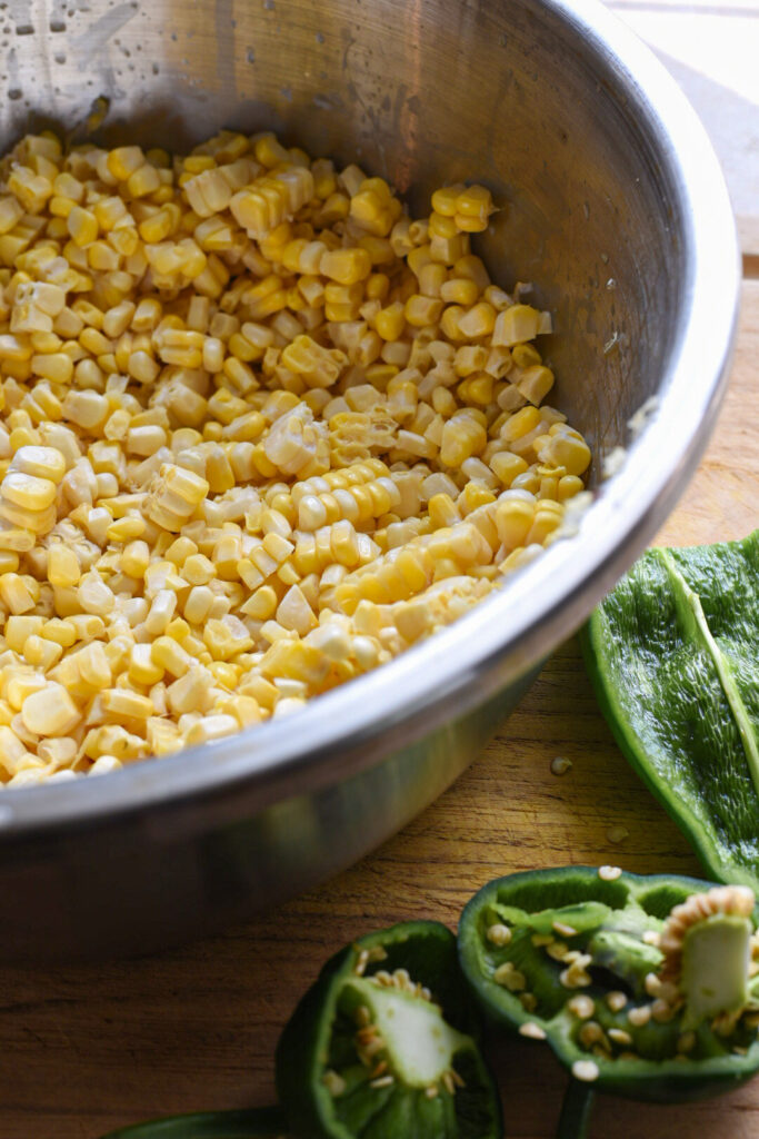 A metal bowl filled with fresh corn kernels and a chopped poblano pepper next to it on a wooden cutting board.