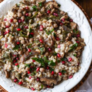 Overhead shot of a large white serving bowl filled with mushroom and barley salad, red pomegranate and green parsley.
