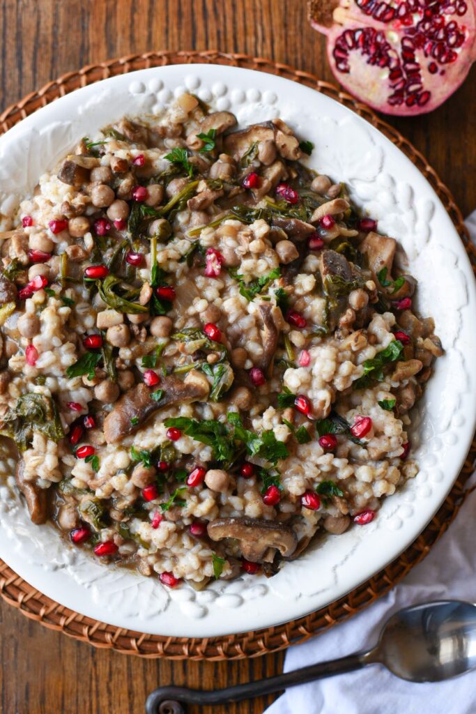 A large white serving bowl filled with barley salad with lemon gravy, topped with bright red pomegranate kernels and green parsley.