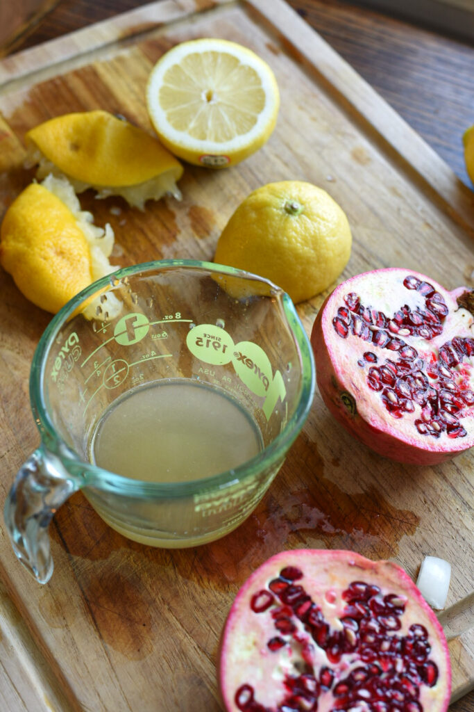 A juiced lemon on a wooden cutting board next to a measuring cup filled with lemon juice and a halved pomegranate.