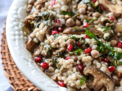 A phot of half of a large white serving bowl filled with barley salad, topped with green parsley and red pomegranate kernels.