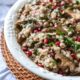 A phot of half of a large white serving bowl filled with barley salad, topped with green parsley and red pomegranate kernels.