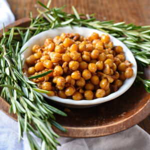 A bowl of rosemary roasted chickpeas and walnuts in a wooden bowl surrounded by fresh rosemary sprigs