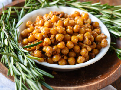 A bowl of rosemary roasted chickpeas and walnuts in a wooden bowl surrounded by fresh rosemary sprigs