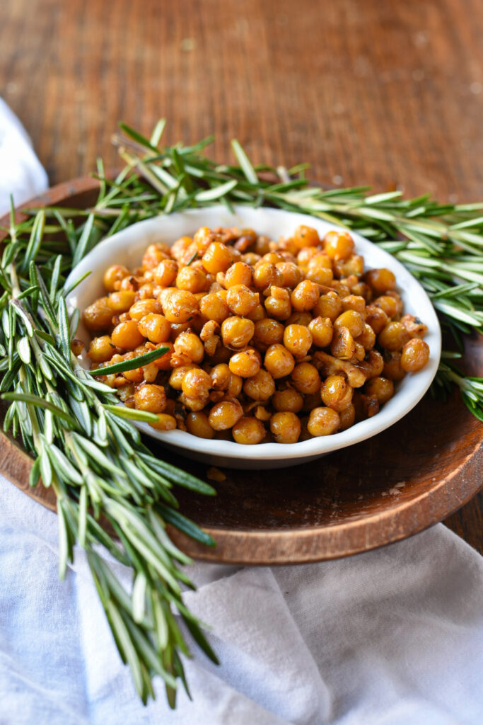 A bowl of rosemary roasted chickpeas and walnuts in a wooden bowl surrounded by fresh rosemary sprigs