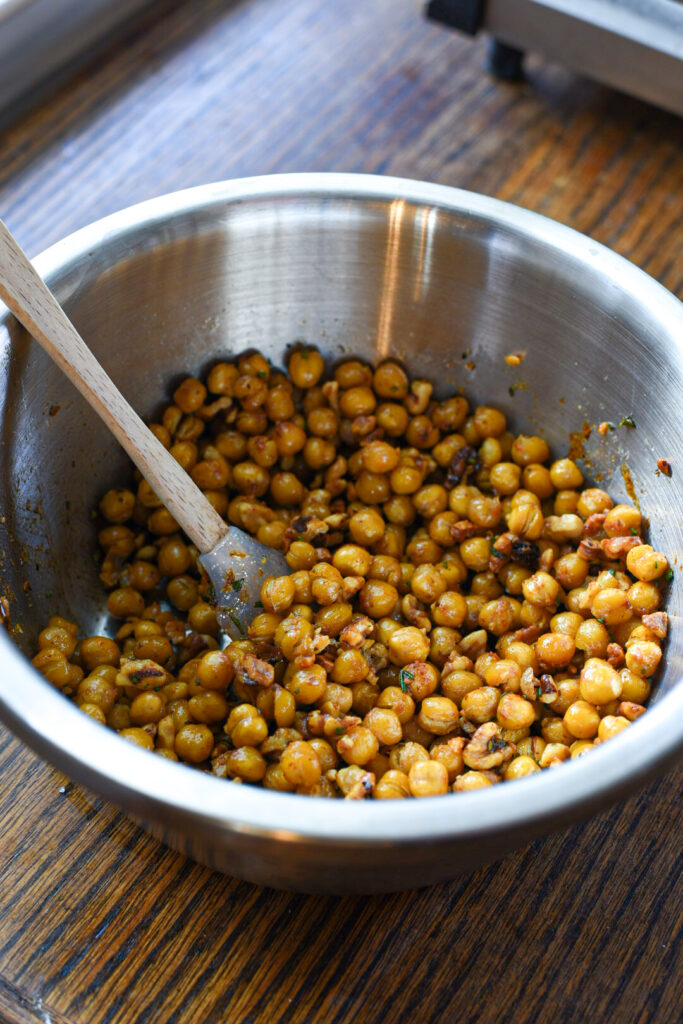 A large metal bowl filled with roasted chickpeas and walnuts with a spoon in the bowl.
