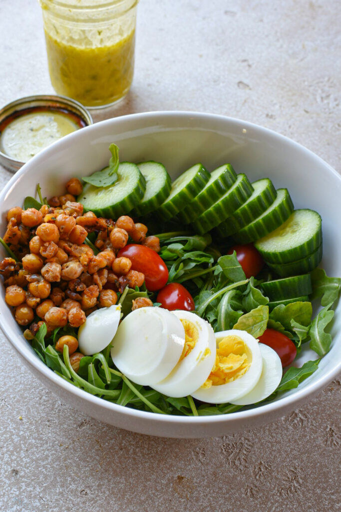 A salad bowl filled with arugula, sliced egg, cucumber, tomatoes and rosemary roasted chickpeas