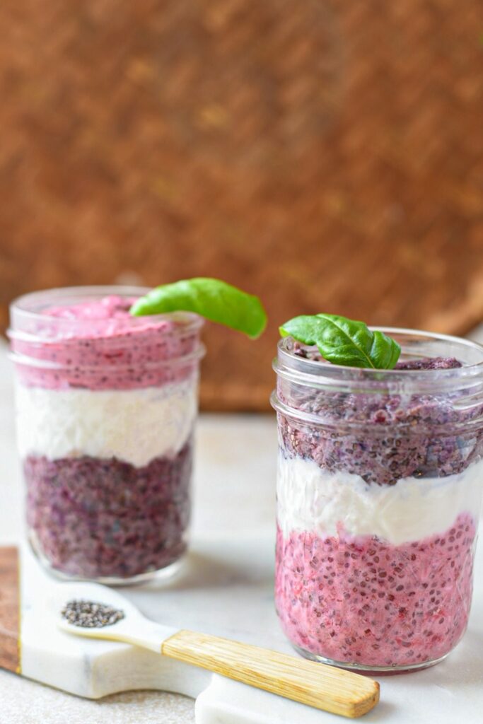 Two glass mason jars filled with red and purple chia seed pudding and white yogurt in a layering pattern