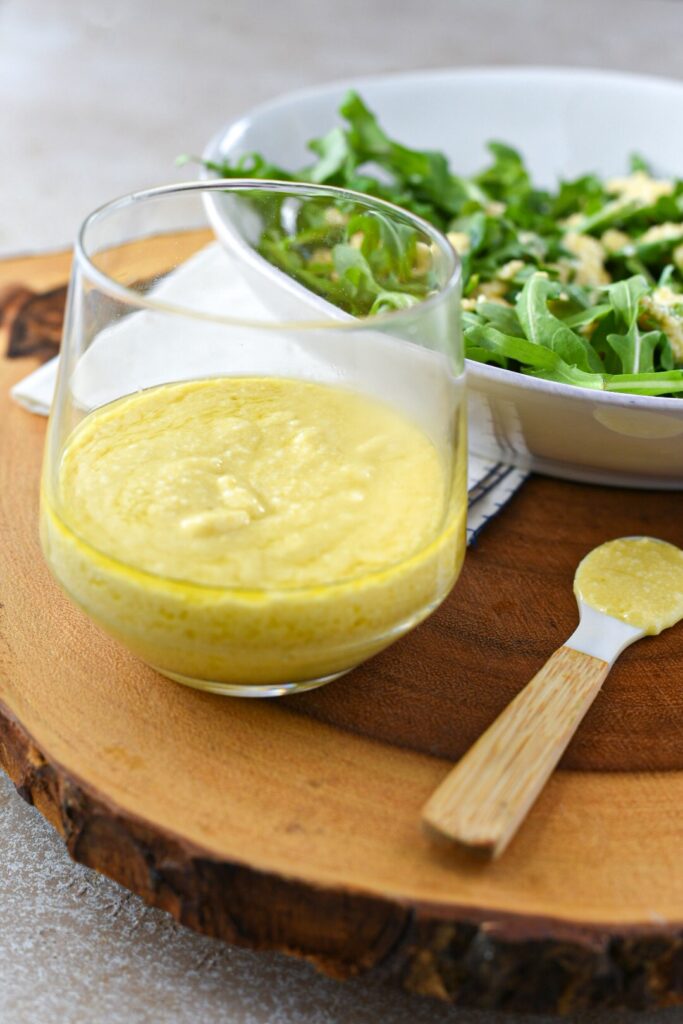 A cup of smooth parmesan vinaigrette on a wooden surface next to an arugula salad