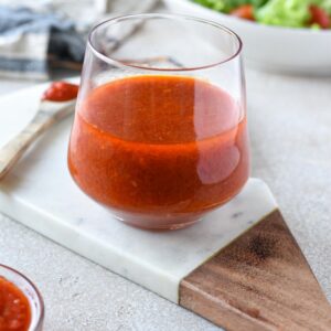 A clear glass filled with bright red harissa vinaigrette with a salad in the background