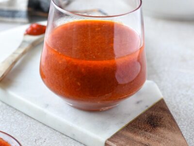 A clear glass filled with bright red harissa vinaigrette with a salad in the background