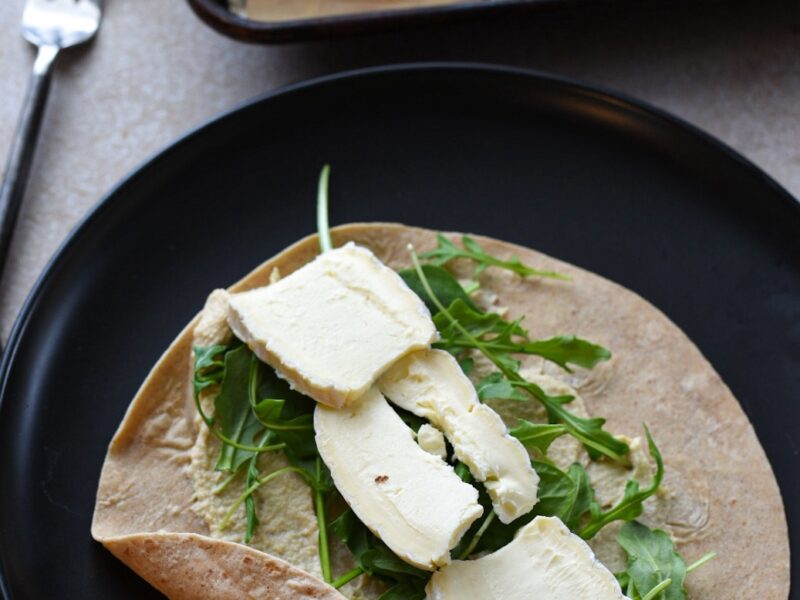 healthy hummus wrap with cheese A wrap on a black plate topped with hummus, spinach and arugula, and brie cheese.