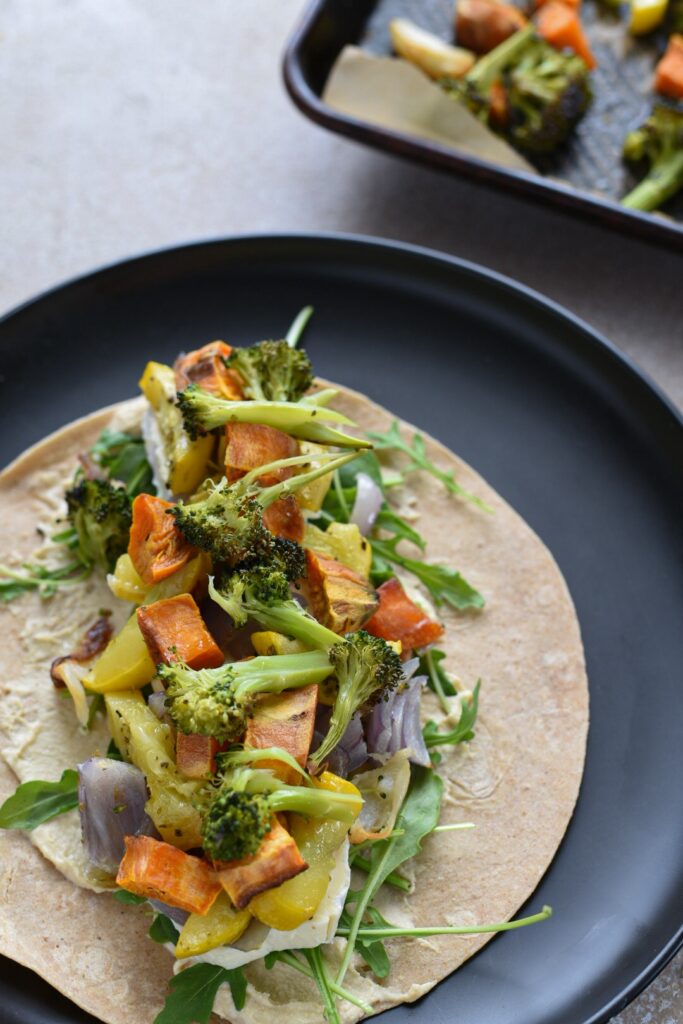 An open tortilla wrap on a black plate filled with roasted veggies including broccoli, sweet potato, yellow squash, red onion, salad greens and hummus.