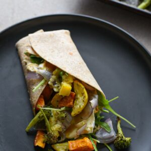 A roasted veggie wrap on a black plate with vegetables spilling out