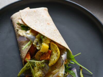 A roasted veggie wrap on a black plate with vegetables spilling out