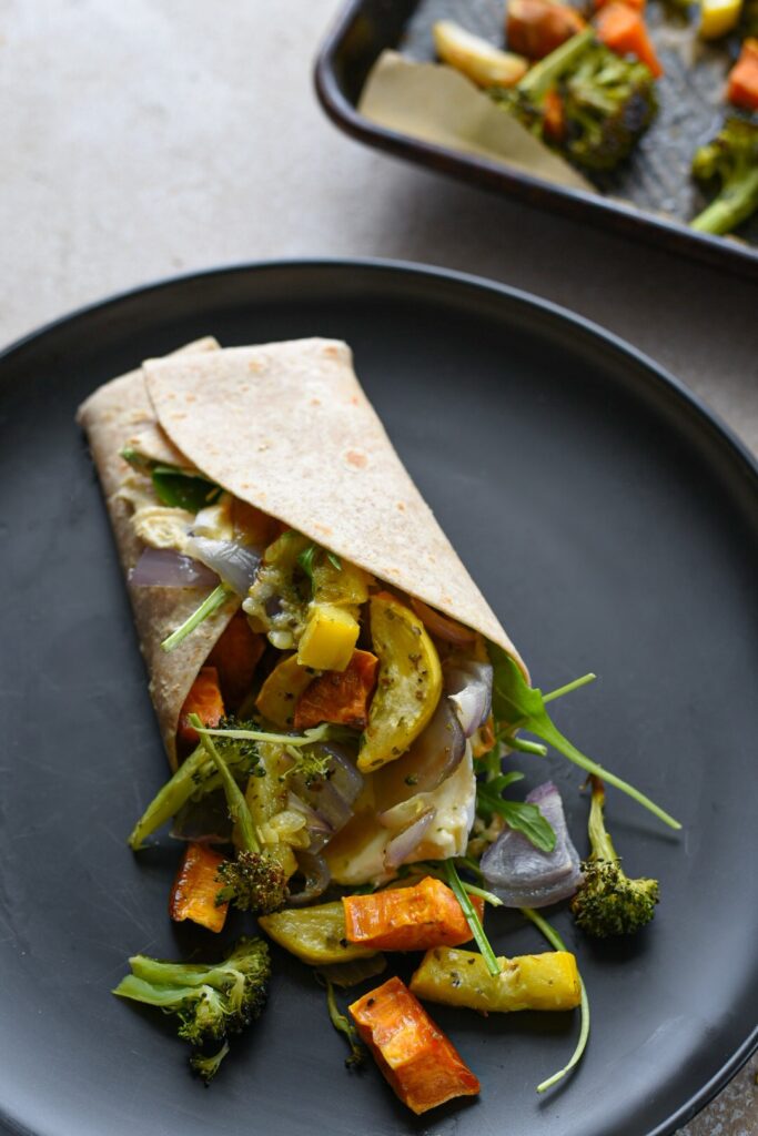 A roasted veggie wrap with hummus on a black plate with roasted vegetables spilling out of it