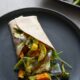 A roasted veggie wrap on a black plate with vegetables spilling out