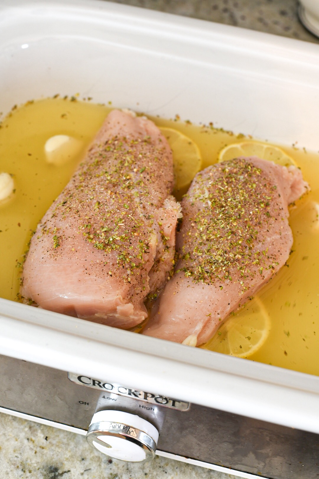 Seasoned chicken breasts on top of lemon slices surrounded by garlic cloves and broth in a slow cooker.
