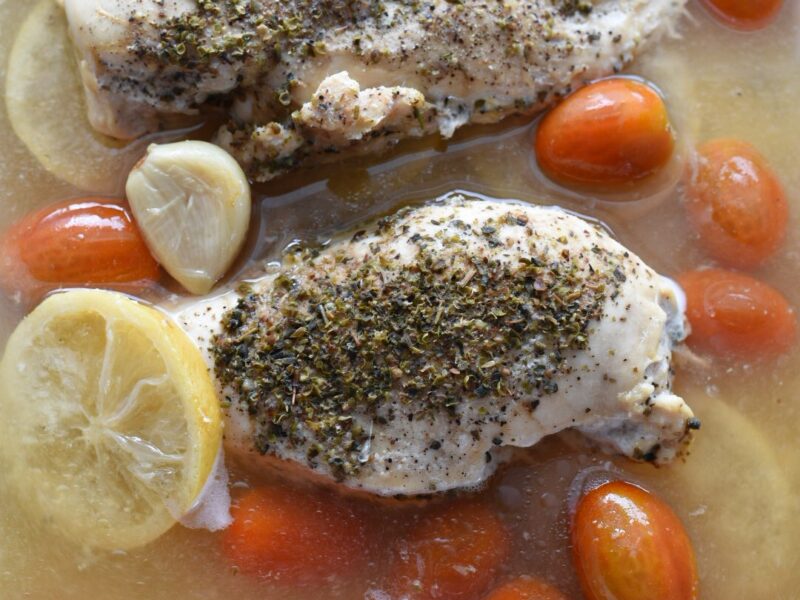slow cooker lemon chicken recipe (2) Two chicken breasts topped with herbs and surrounded by broth, lemon slices and cherry tomatoes in a slow cooker.