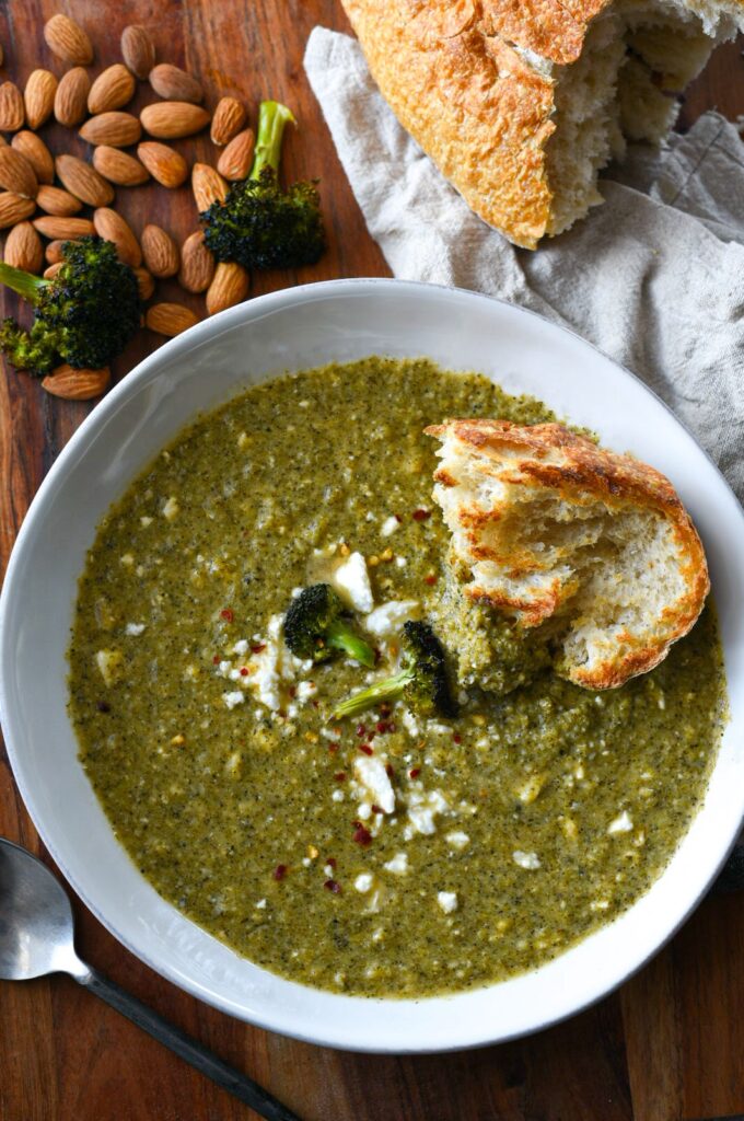Broccoli feta soup in a white bowl with almonds on the table and bread in the soup
