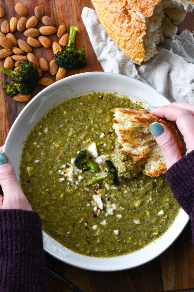 Hands dipping toasted bread into broccoli feta soup with almonds