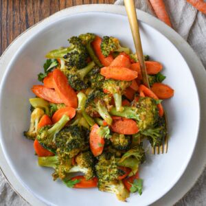 An overhead shot of a bowl filled with roasted broccoli and carrots with raw carrots in the corner