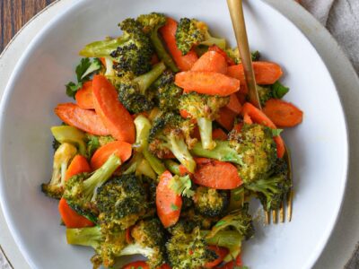 An overhead shot of a bowl filled with roasted broccoli and carrots with raw carrots in the corner