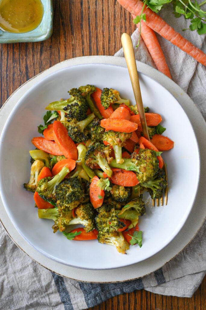 An overhead shot of a bowl filled with roasted broccoli and carrots with raw carrots in the corner