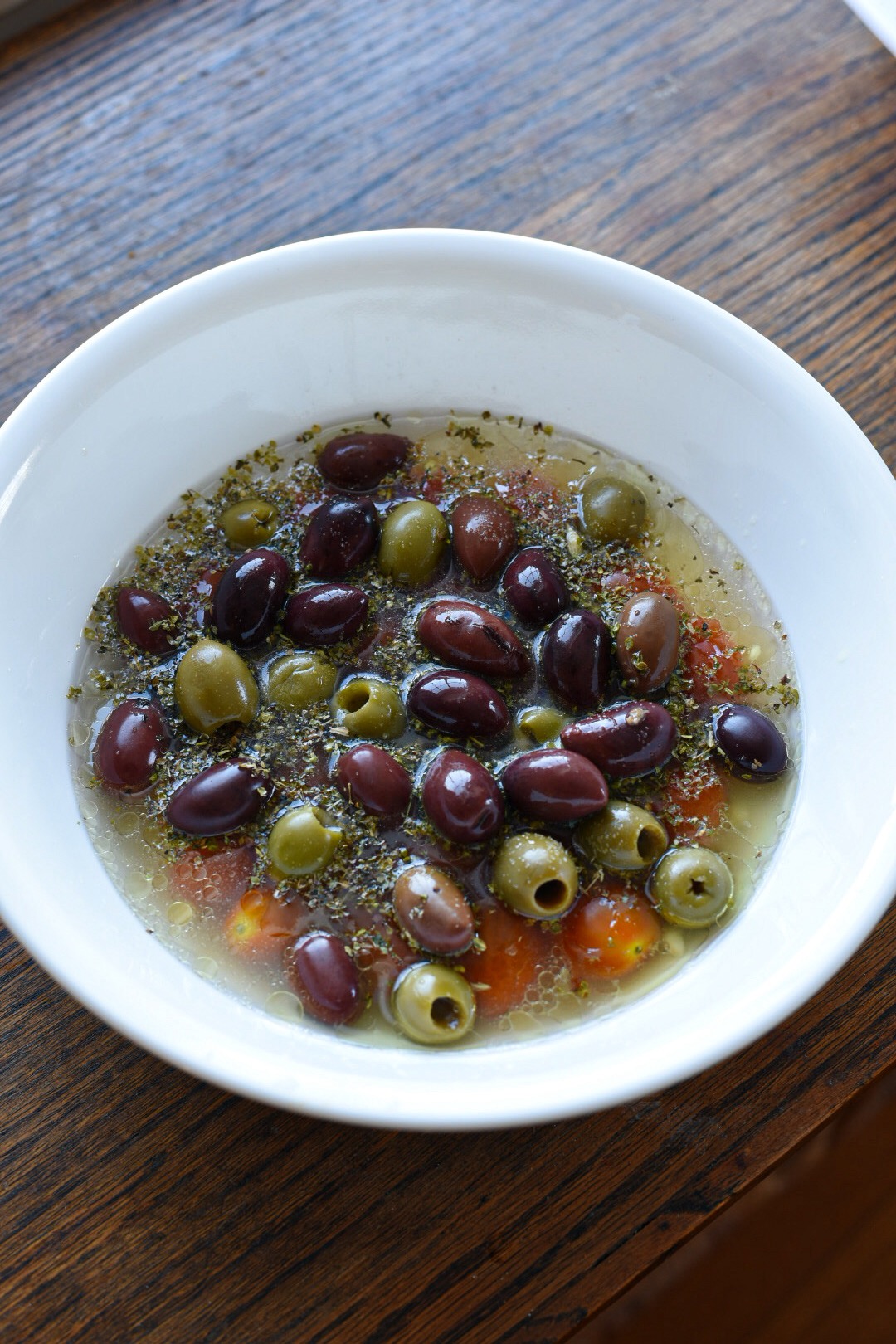 Green and kalamata olives in a white bowl with cherry tomatoes, italian seasoning, broth, lemon juice and white wine.