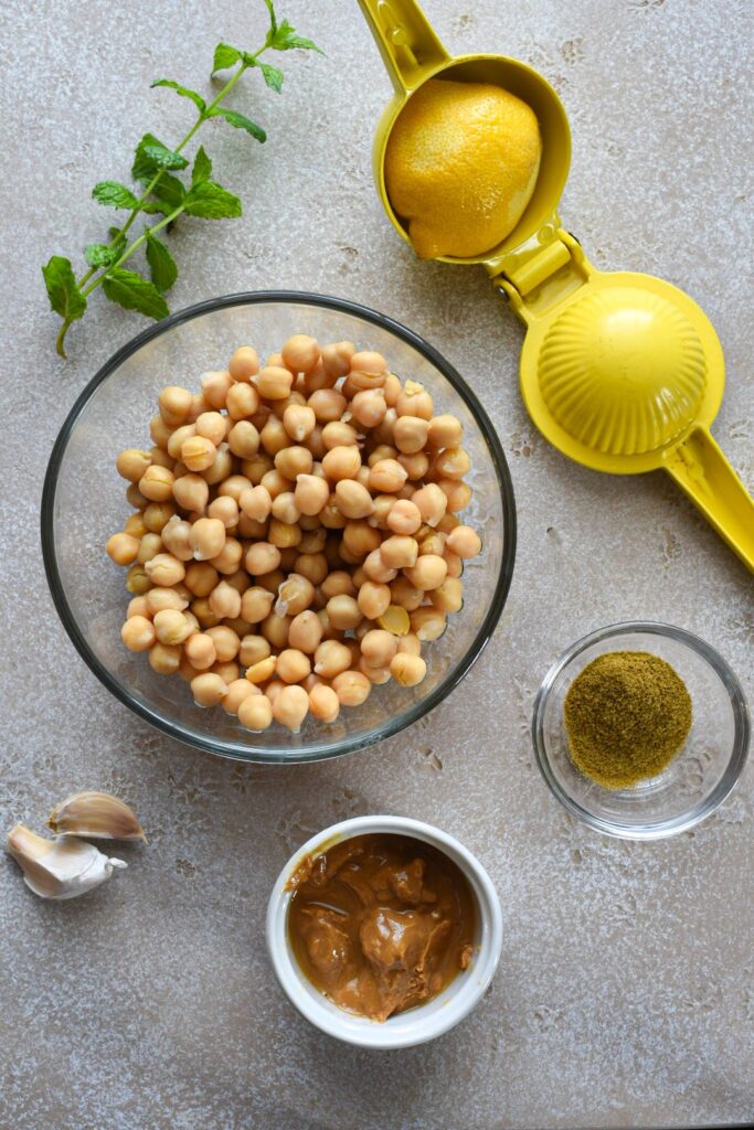 An overhead shot of the ingredients used to make homemade hummus without tahini, including chickpeas, cumin, mint leaves, garlic, peanut butter and a lemon.