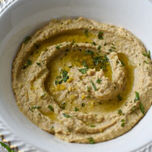 A white bowl with gold trip filled with a hummus recipe without tahini topped with olive oil and mint.