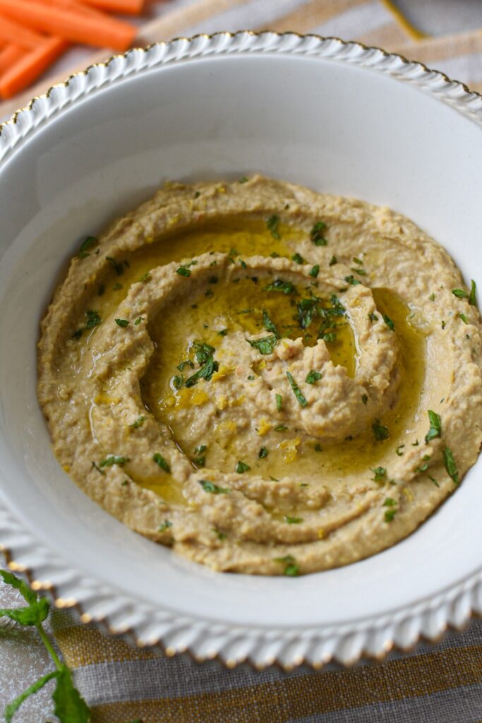 A white bowl with gold trip filled with a hummus recipe without tahini topped with olive oil and mint.