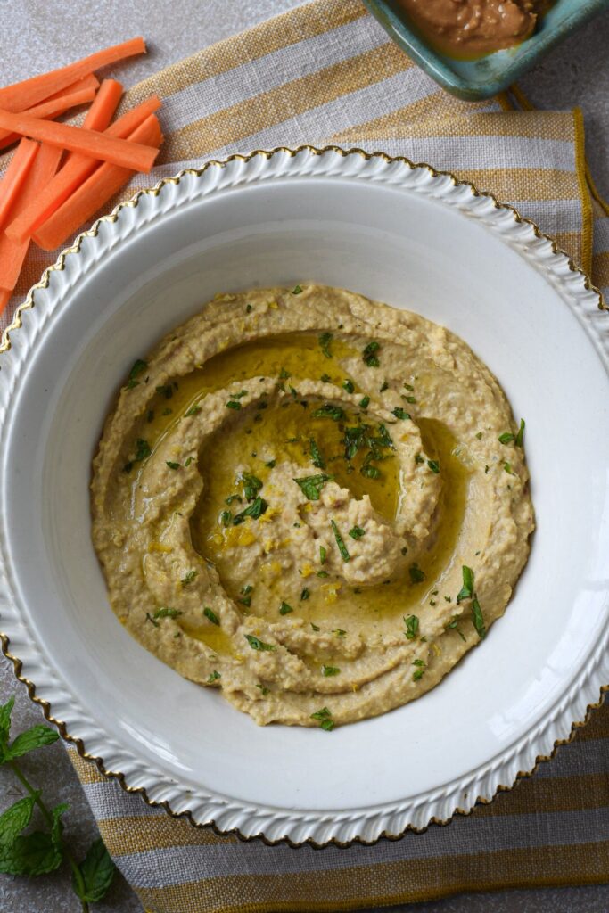 An overhead shot of hummus without tahini in a white bowl with carrot sticks next to it and  minced mint leaves on top.