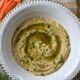 An overhead shot of hummus without tahini in a white bowl with carrot sticks next to it and minced mint leaves on top.