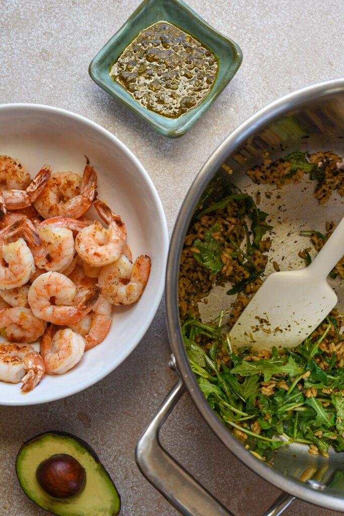 Cooked shrimp in a white bowl next to arugula and grains in another large pot.