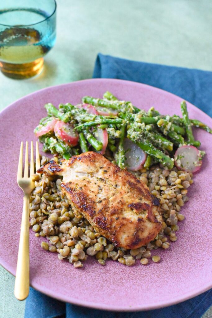 A pink plate with pan a cooked chicken breast, lentils and blanched asparagus salad with gremolata sauce