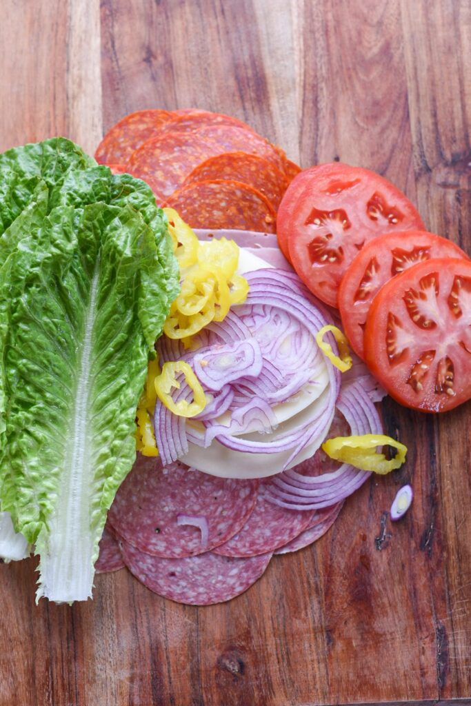 The ingredients for a chopped italian sandwich on a cutting board, including meat, cheese, lettuce, tomatoes, and red onions.