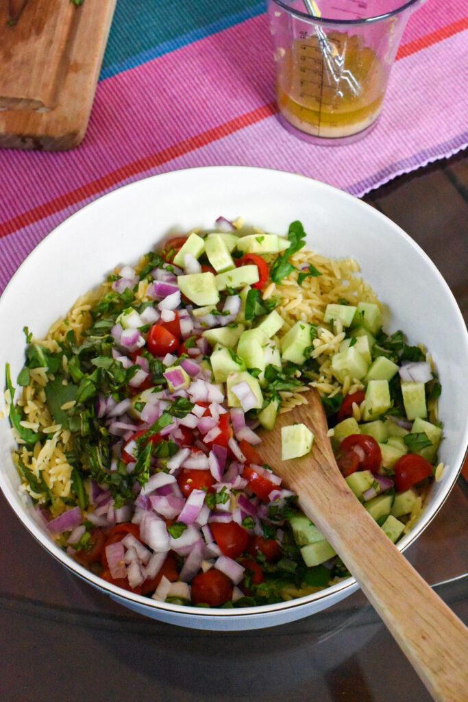 easy orzo pasta salad recipe A large serving bowl of orzo topped with chopped red onion, cherry tomatoes, cucumber, arugula and basil.