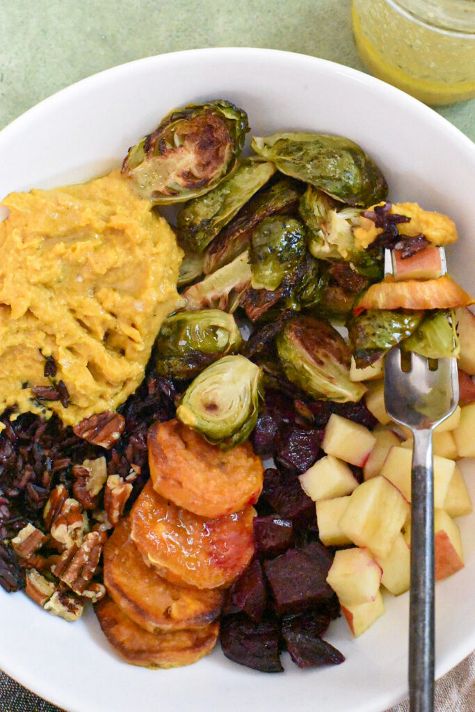 A fall harvest bowl with a fork with sweet potato, brussels sprouts, pumpkin hummus, beets, apple, walnuts, wild rice and dressing.