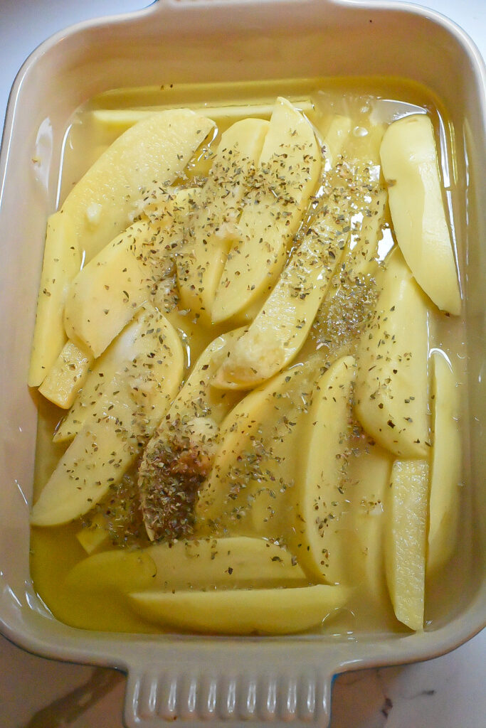Wedged potatoes in a baking dish topped with lemon juice, broth, olive oil and oregano for greek potatoes.