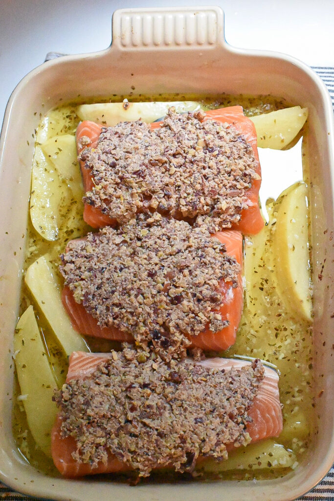 Three raw salmon filets topped with a walnut crust over greek potatoes in a baking dish.
