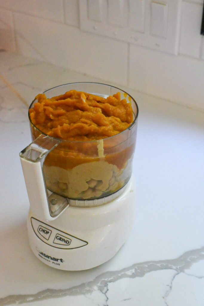 Ingredients for pumpkin hummus in a small food processor on a white countertop.