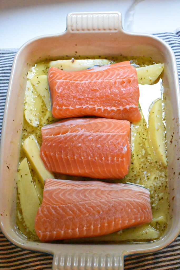 Three raw salmon filets over greek potatoes in a baking dish.