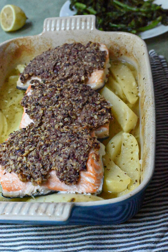 Three pieces of baked walnut crusted salmon over greek potatoes in a blue baking dish.
