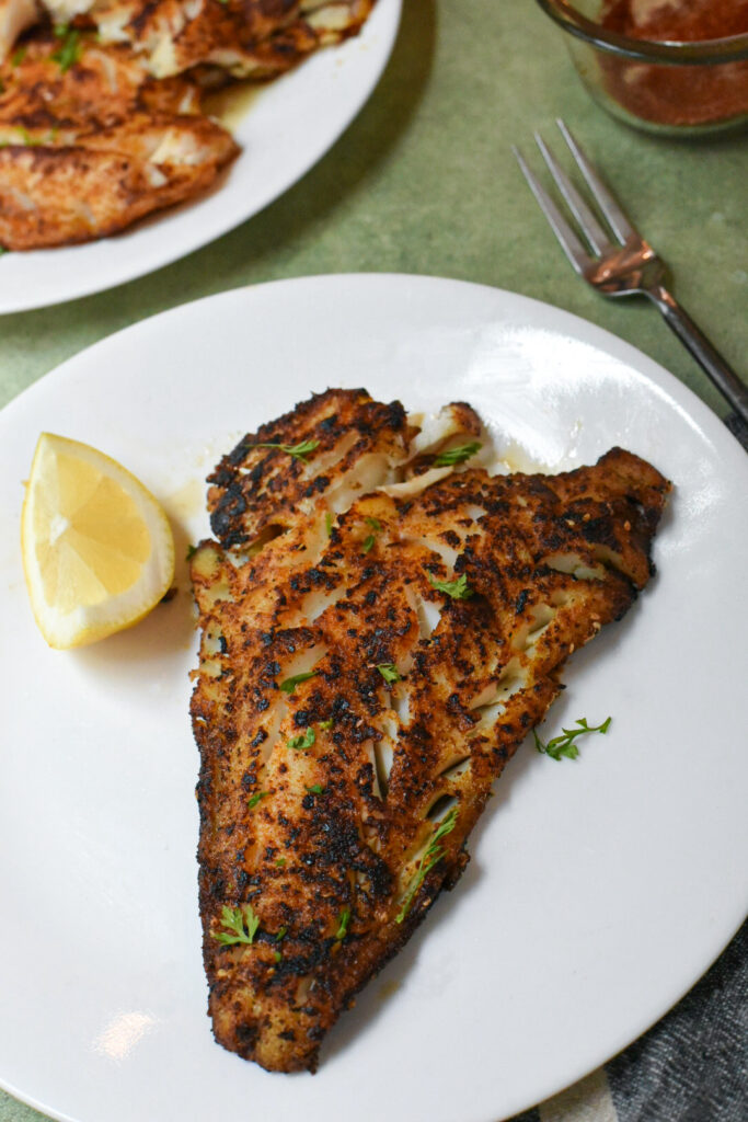 A blackened cod filet on a white plate with a lemon slice next to it.