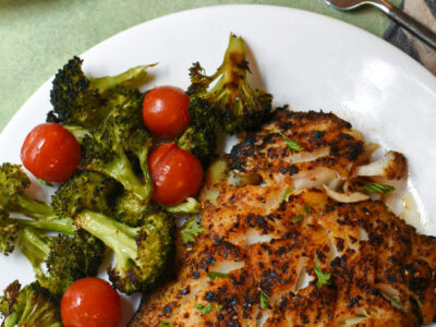A plate with blackened cod and broccoli and tomatoes over a green surface.