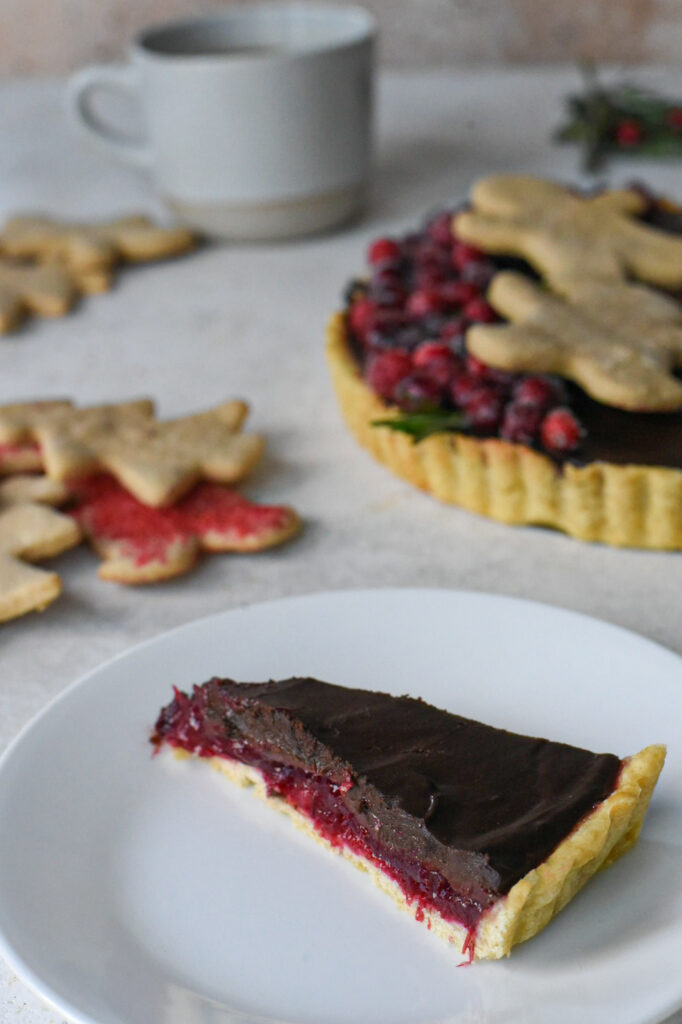 A slice of chocolate cranberry tart on a white plate with christmas cookies behind it.