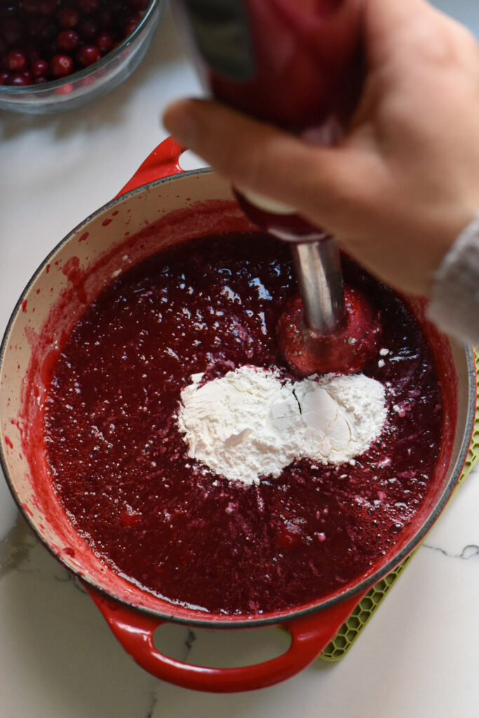 A dutch oven filled with the makings of a cranberry filling for a tart, with an immersion blender blending cornstarch into it.