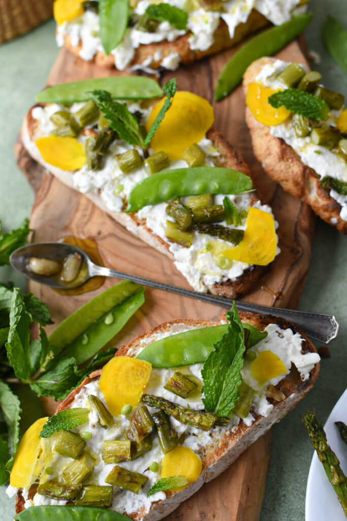 Burrata toast on a wooden serving platter with garlic confit and spring vegetables.